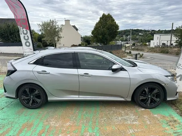 Vue de profil côté gauche d'une Honda Civic grise 5 portes avec jantes noires et vitres teintées, stationnée en extérieur.