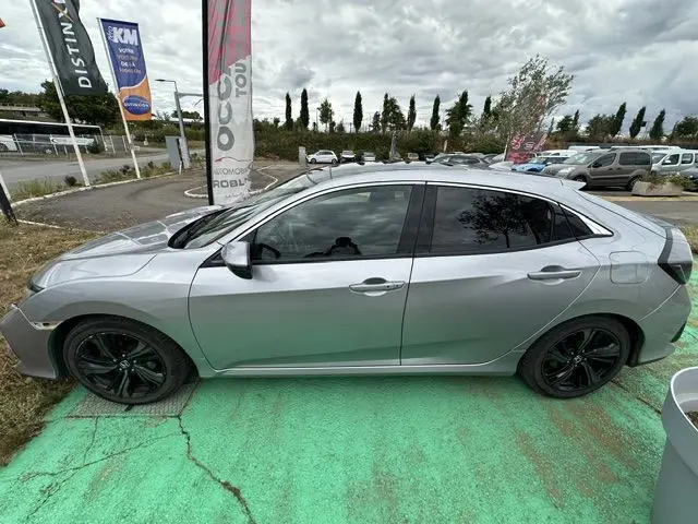 Vue de profil côté gauche d'une Honda Civic grise 5 portes avec jantes noires, stationnée sur un sol vert.