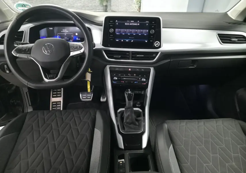 Vue intérieure avant du Volkswagen T-Roc 2025 noir, avec volant multifonction, écran tactile central et sièges tissu noir à motifs géométriques.