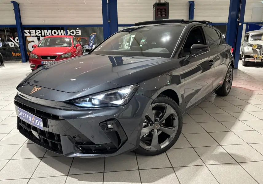 Vue 3/4 avant d'une CUPRA Leon gris Graphène avec jantes anthracite et signature lumineuse Tri-LED visible en intérieur showroom.