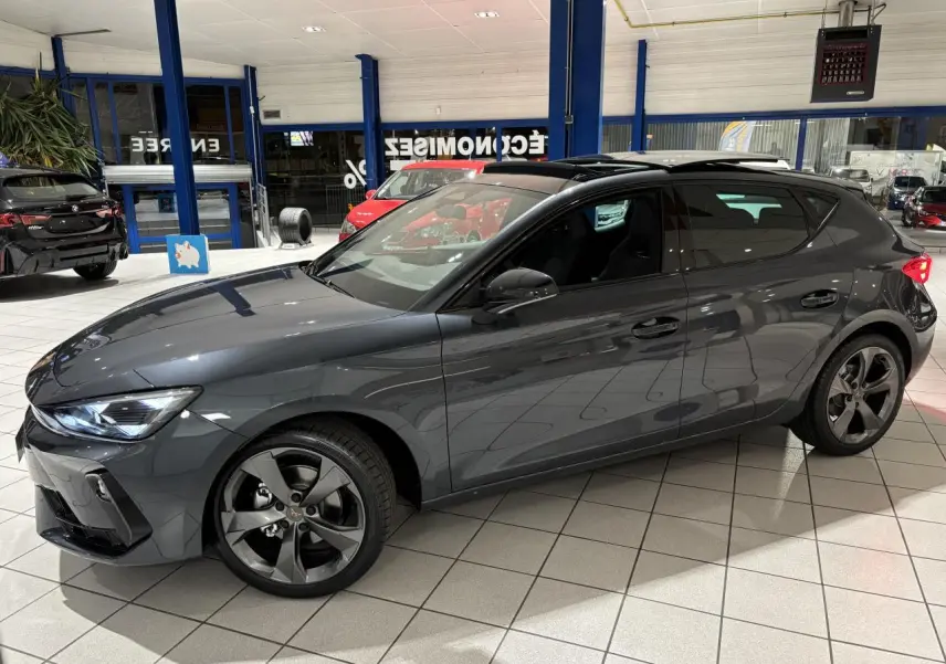 Vue de profil côté gauche d’une CUPRA Leon gris Graphène avec jantes anthracite en showroom lumineux.