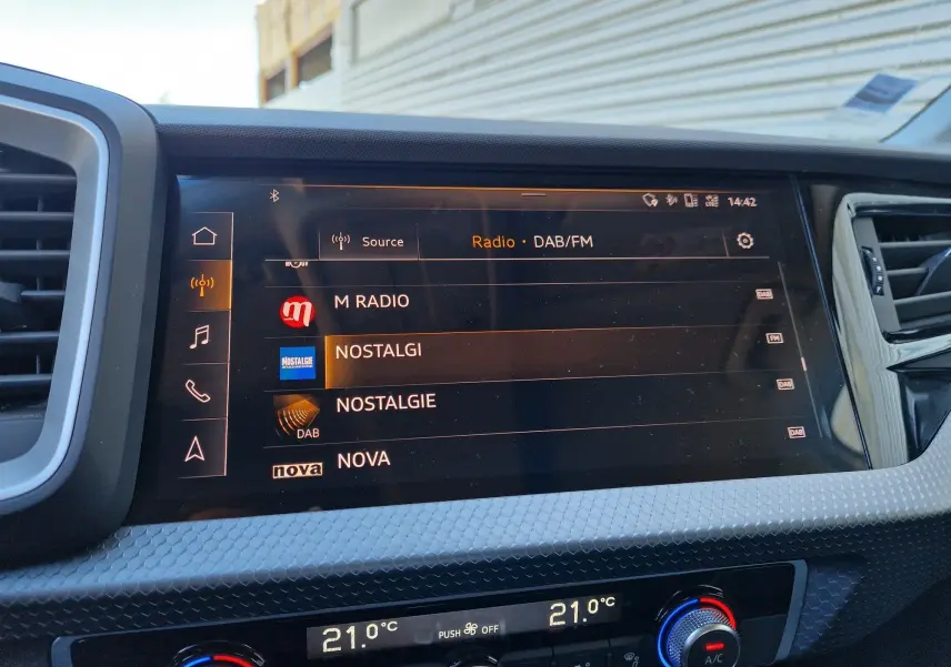 Vue rapprochée de l’écran multimédia et de la console de climatisation de l’Audi A1 Sportback blanc glacier, affichant les radios DAB/FM.
