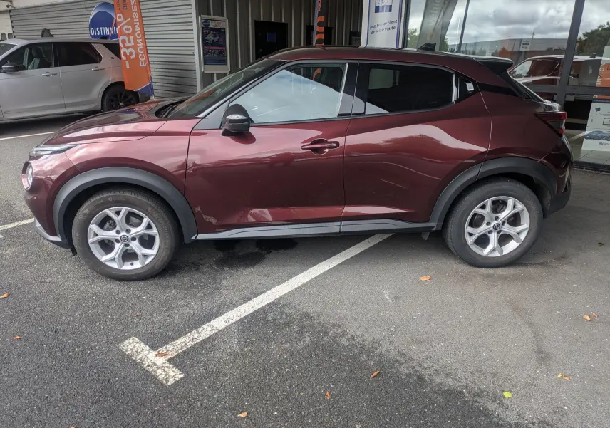 Vue de profil côté gauche d'un Nissan Juke rouge 2020, soulignant ses jantes alliage et ses protections noires sur les passages de roue.