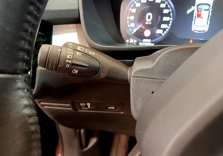Gros plan sur le commodo d’éclairage à gauche du volant noir, avec tableau de bord digital du Volvo XC40 T2 2020 en arrière-plan.