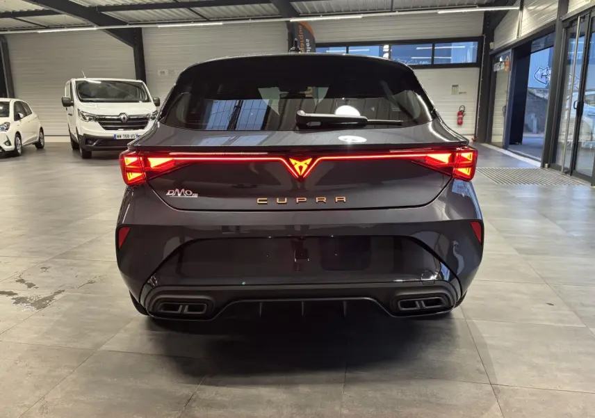 Vue arrière d'une CUPRA Leon gris magnetic avec feux arrière Tri-LED et double sortie d'échappement noire en intérieur showroom.