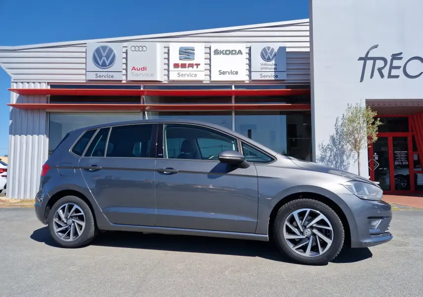 Vue de profil côté gauche d'une Volkswagen Golf Sportsvan gris Indium Metal stationnée devant un garage multimarques.