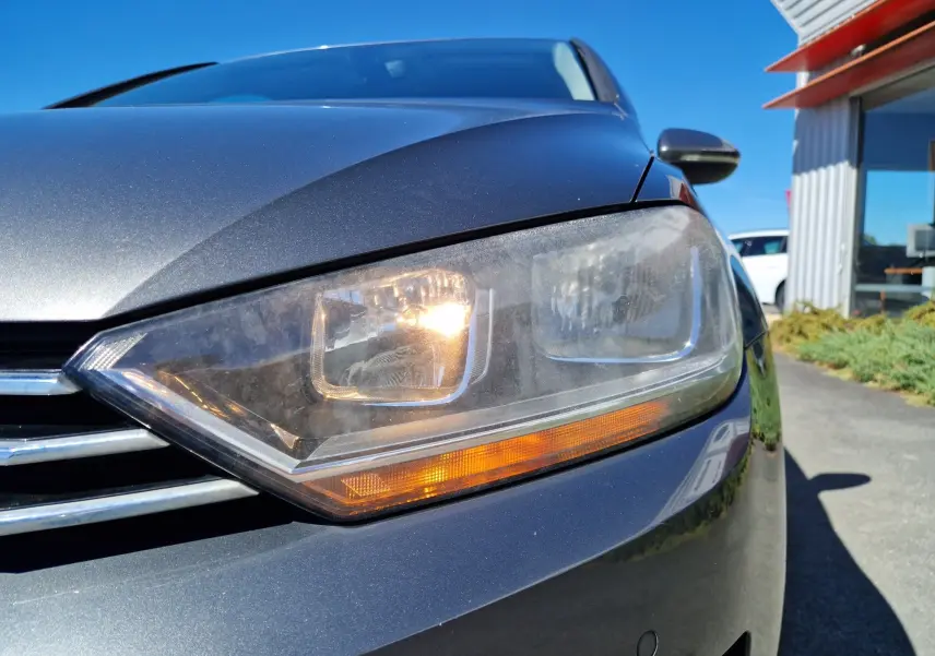 Gros plan sur le phare avant droit allumé d'une Volkswagen Golf Sportsvan gris Indium Metal sous un ciel bleu.