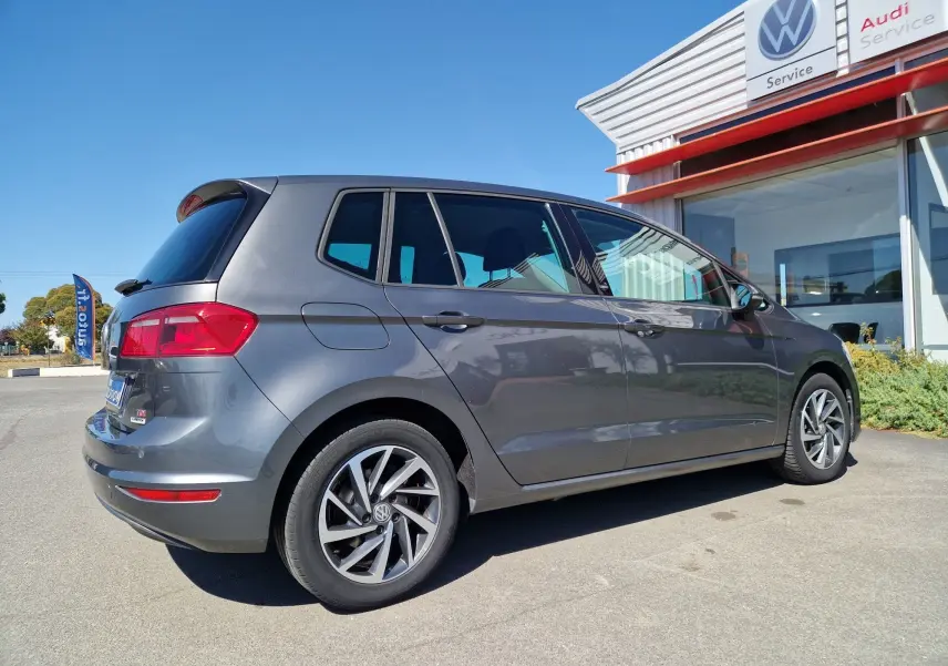Volkswagen Golf Sportsvan gris Indium métal vue 3/4 arrière côté droit, stationnée devant un service Volkswagen.