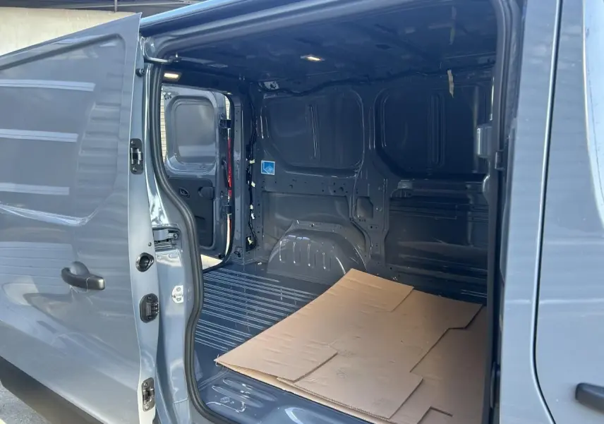 Vue intérieure du Renault Trafic Fourgon gris urbain, porte latérale ouverte montrant l'espace de chargement vide avec carton au sol.