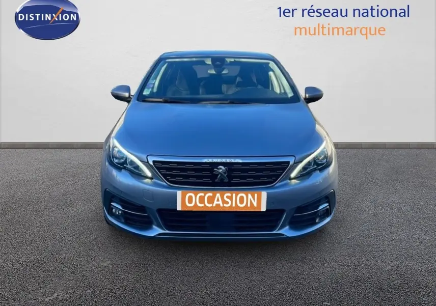 Vue frontale d'une Peugeot 308 gris Artense métal de 2019 avec phares allumés et plaque "OCCASION" visible.