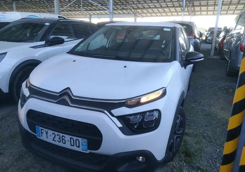 Vue 3/4 avant d'une Citroën C3 Société blanche de 2021 avec feux allumés, garée sous abri sur sol gravillonné.