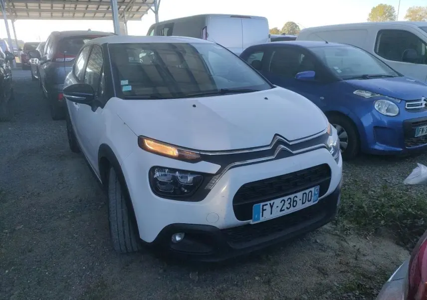Vue 3/4 avant d'une Citroën C3 Société blanche de 2021 avec feux allumés, garée sur un sol en gravier.