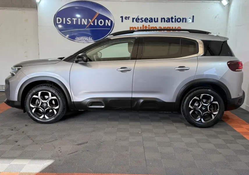 Vue de profil côté gauche d’un Citroën C5 Aircross gris clair avec jantes noires et toit noir en intérieur showroom.