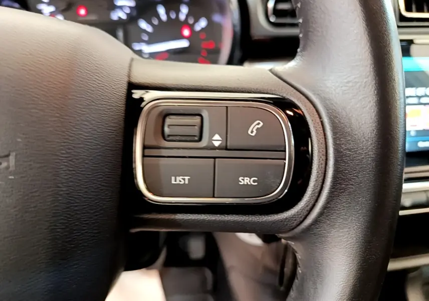 Gros plan sur les commandes gauche du volant cuir d’une Citroën C3 blanche, avec tableau de bord flou en arrière-plan.