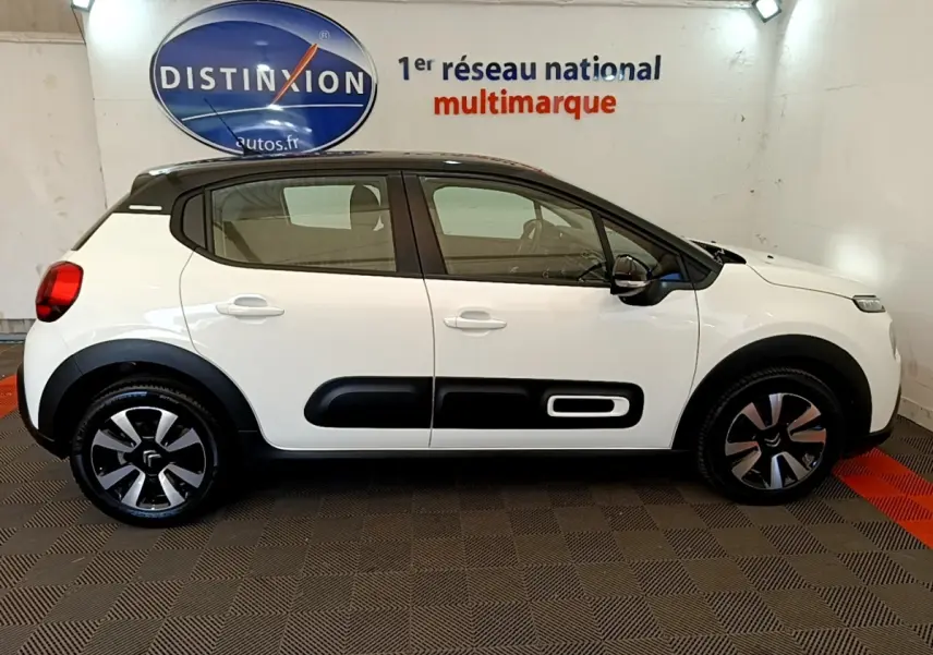 Profil latéral droit d'une Citroën C3 blanche avec toit noir et protections latérales noires, en intérieur showroom.