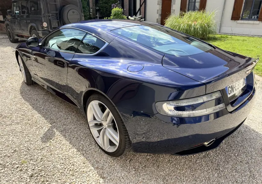 Vue 3/4 arrière droite d'une Aston Martin DB9 5.9 V12 bleu foncé brillant, avec jantes argentées et feux arrière LED.