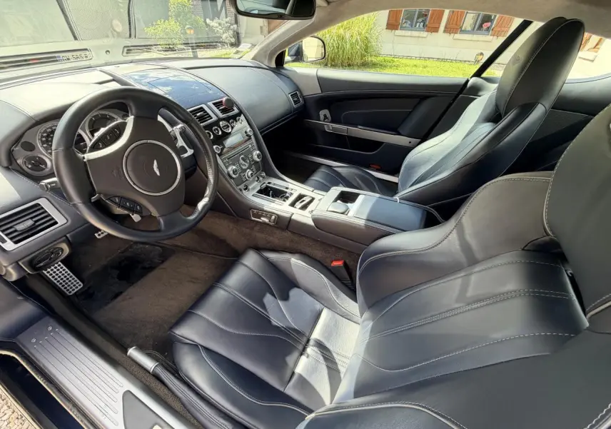 Vue intérieure côté conducteur de l'Aston Martin DB9 2016, sièges cuir noir avec coutures contrastées et tableau de bord épuré.