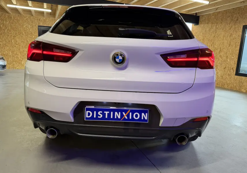 Vue arrière d'une BMW X2 sDrive18d blanche avec double sortie d'échappement chromée et feux arrière LED allumés.