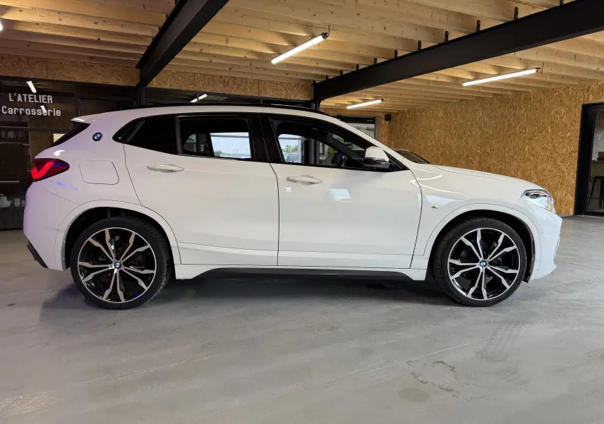 BMW X2 sDrive18d blanc vue de profil droit en intérieur, avec jantes alu 19'' et toit panoramique visible.