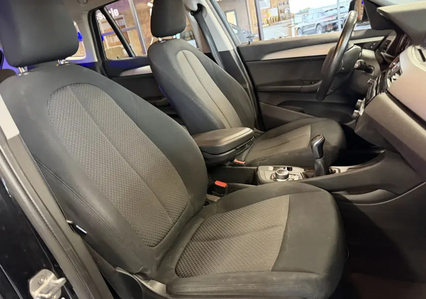 Vue intérieure côté conducteur de la BMW X1 noire 2019, montrant les sièges avant en tissu et la console centrale avec levier de vitesses.