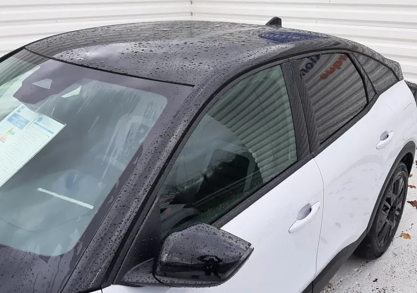 Vue 3/4 avant droit de la Citroën C4 hybride blanche Okenite avec toit et rétroviseur noir brillant sous pluie.