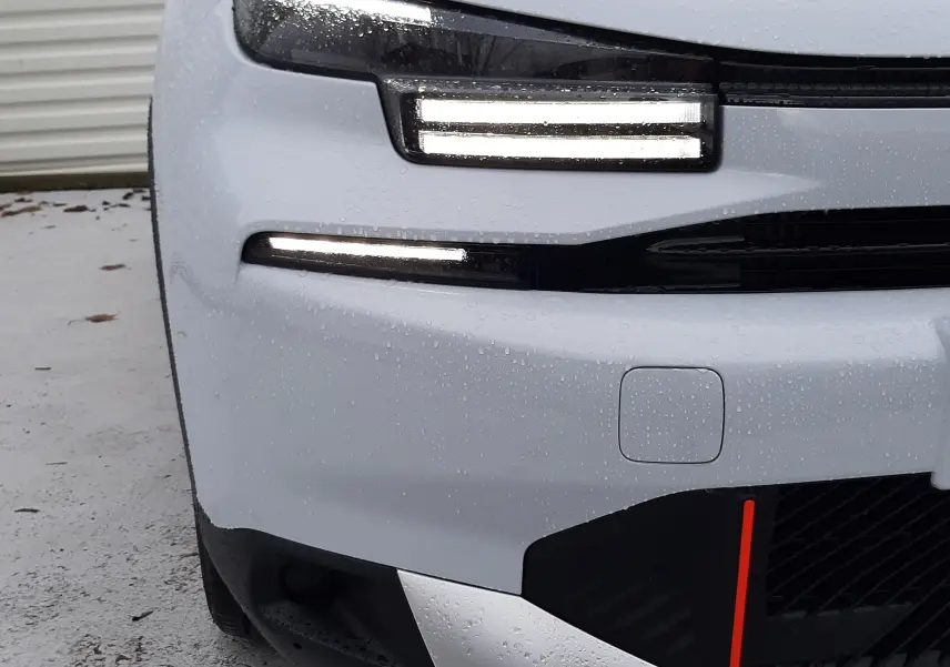 Gros plan sur l'avant gauche blanc Okenite d'une Citroën C4 1.2 Hybrid 145ch avec phares LED allumés sous la pluie.