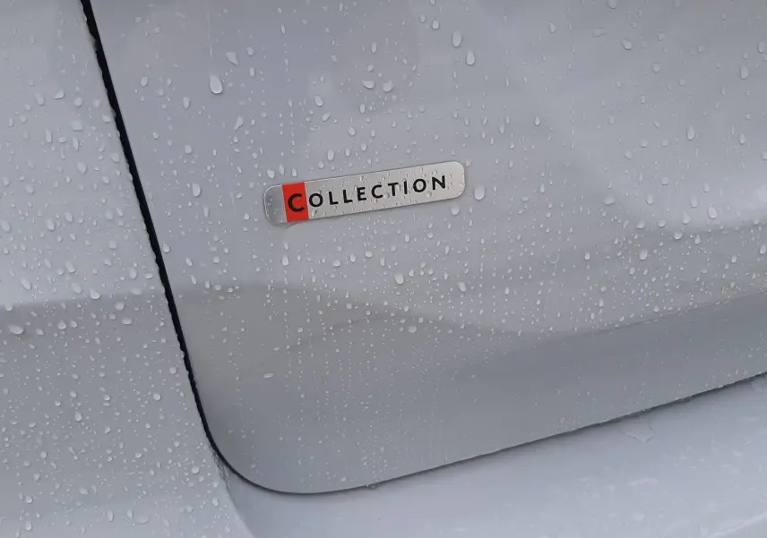 Gros plan sur l'écusson "Collection" sur la porte arrière blanche Okenite d'une Citroën C4, avec gouttes de pluie visibles.