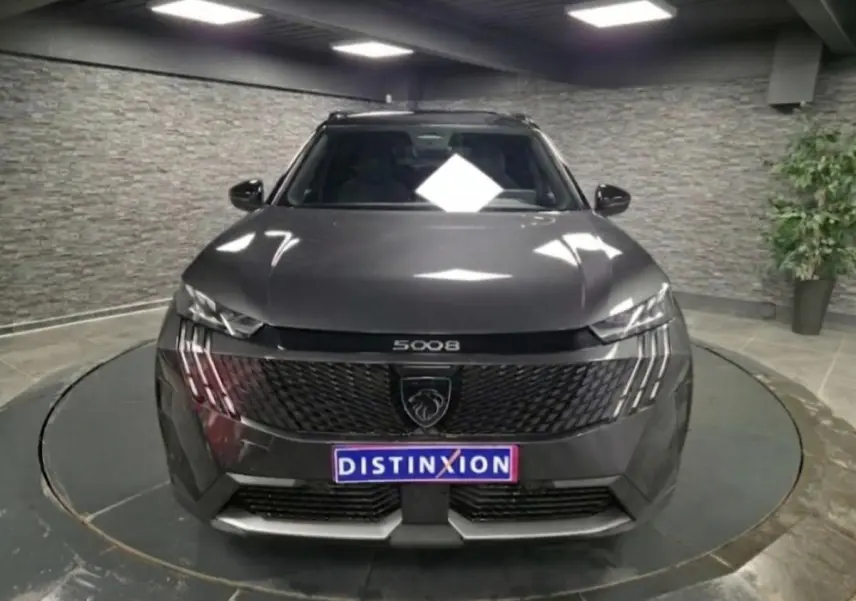 Vue avant d'un Peugeot 5008 gris métallisé 2025 avec calandre noire et feux LED à trois griffes, en intérieur.