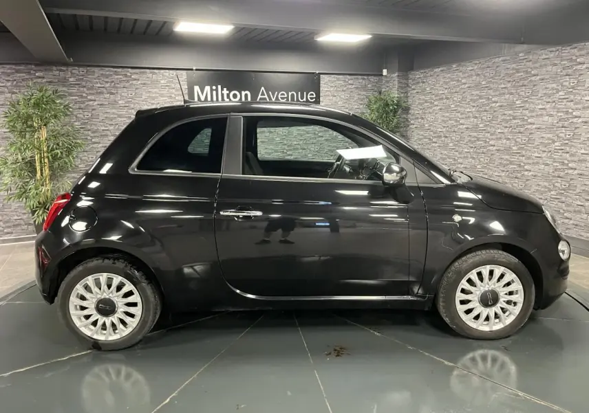 Profil droit de la Fiat 500 noire 2023 avec jantes alliage blanches et rétroviseur chromé en intérieur showroom.