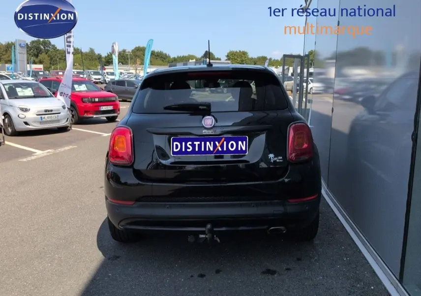Vue arrière d'une Fiat 500X noire Cinéma pastel avec attelage, stationnée près d'un bâtiment vitré.