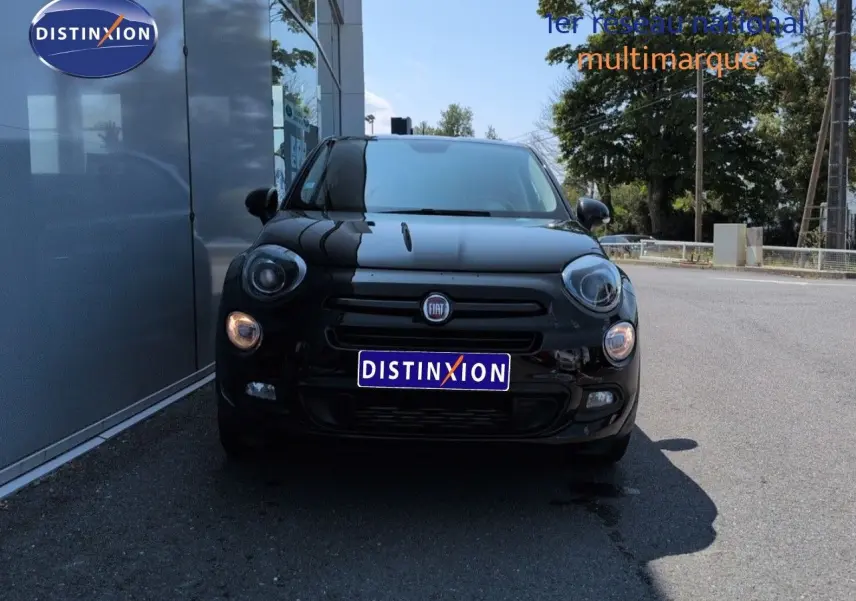 Vue de face d'une FIAT 500X noire Cinéma pastel extra-série, avec phares allumés, stationnée devant un bâtiment.