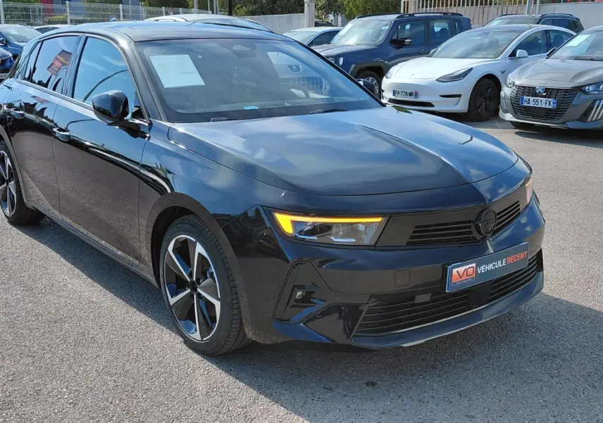 Opel Astra noire 2025 vue 3/4 avant droit avec feux diurnes allumés sur parking extérieur.