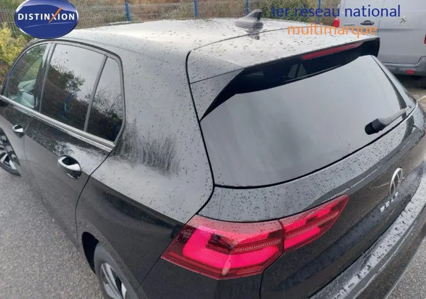 Vue 3/4 arrière droite d'une Volkswagen Golf noire métal 2024 avec feux arrière allumés sous la pluie.
