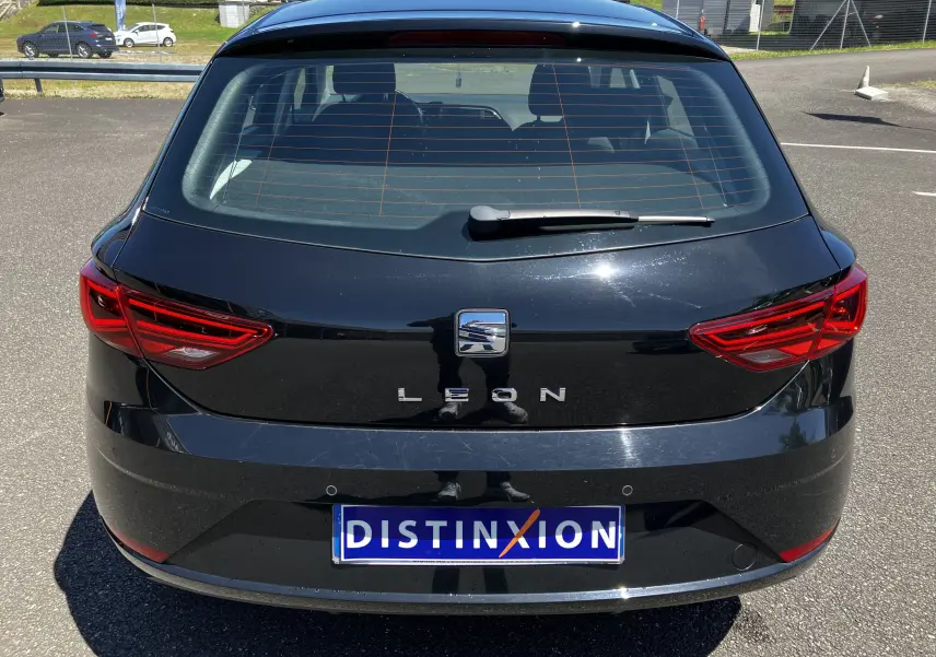 Vue arrière d'une SEAT Leon noire 5 portes avec feux rouges et logo distinctif sur un parking ensoleillé.