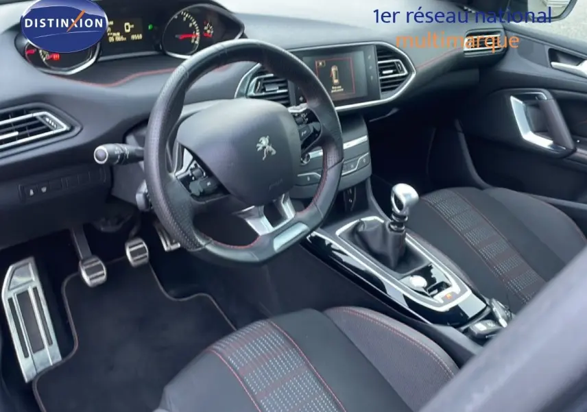 Vue intérieure côté conducteur de la Peugeot 308 gris Artense de 2017 avec volant cuir, boîte manuelle et sièges tissu noir à surpiqûres rouges.