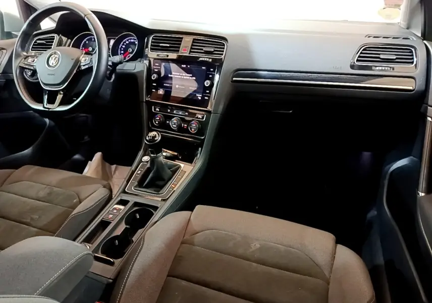 Intérieur de Volkswagen Golf gris clair, vue de face côté conducteur, volant cuir, écran tactile et boîte manuelle visible.