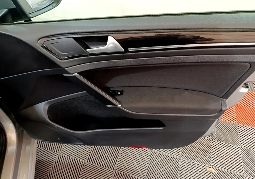 Vue rapprochée de la porte avant gauche d'une Volkswagen Golf gris clair, avec garniture noire et poignée argentée.