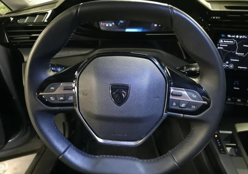 Vue rapprochée du volant cuir noir avec logo Peugeot et commandes intégrées, tableau de bord numérique en arrière-plan.