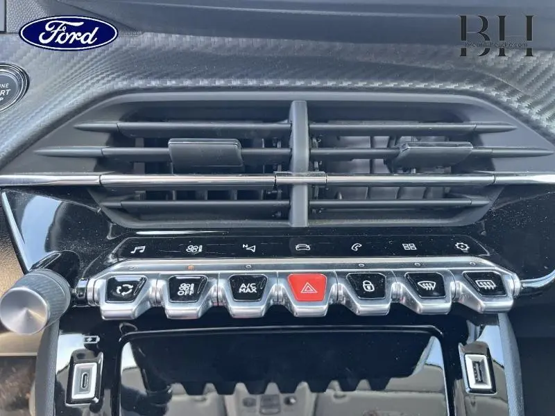 Gros plan sur la console centrale noire brillante de la Peugeot 208 blanche, avec boutons de commande et aérateurs.