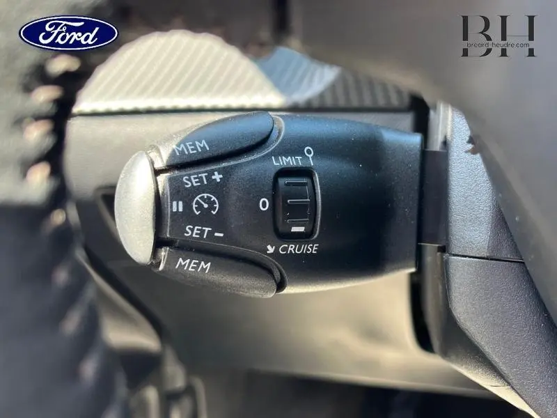 Le véhicule visible est une Peugeot 208 1.5 BlueHDi 100ch S&S Allure Business Blanc Banquise, mais l'image montre un gros plan sur la commande de régulateur de vitesse située sur la colonne de direction,