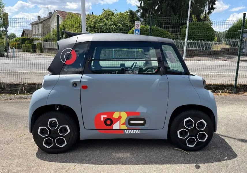 Vue de profil côté gauche d'une Citroën Ami MY AMI POP 2024 gris clair avec toit noir et décor graphique rouge et jaune sur la porte.