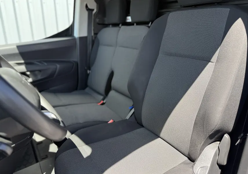 Vue intérieure côté conducteur du Citroën Berlingo XL 2022, sièges tissu gris noir avec accoudoirs et ceintures visibles.