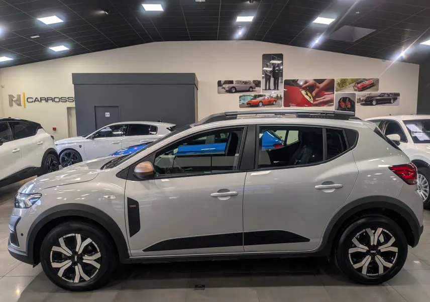 Vue de profil droit d'une Dacia Sandero Stepway 2023 gris Moonstone avec jantes bi-ton et barres de toit noires.