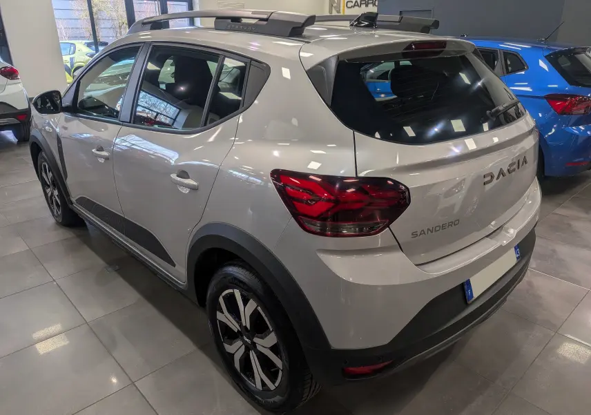 Vue 3/4 arrière droite d'une Dacia Sandero Stepway 2023 gris Moonstone avec jantes biton et barres de toit visibles en showroom.