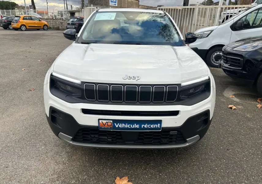 Vue de face d’un Jeep Avenger blanc 2024 avec calandre noire et phares LED intégrés, sur parking extérieur.