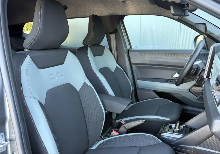 Vue intérieure côté conducteur du Dacia Duster 2026, sièges bicolores gris clair et noir avec accoudoir central.
