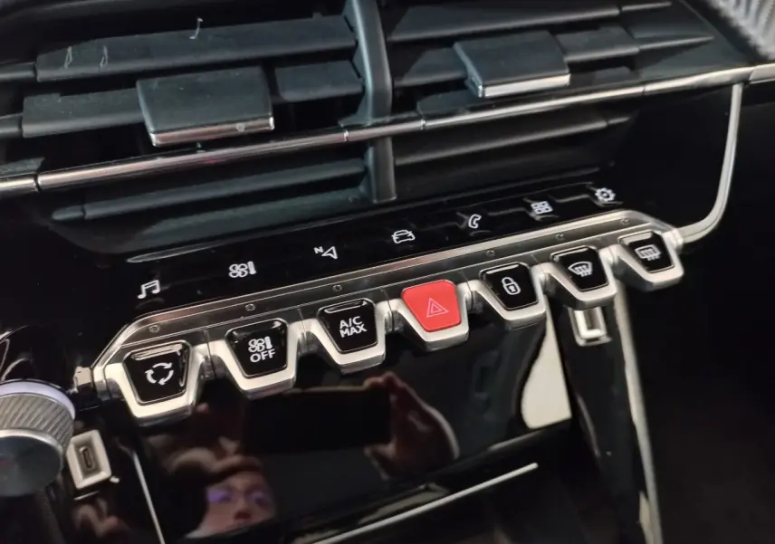 Gros plan sur la rangée de boutons chromés et noirs du tableau de bord du Peugeot 2008 orange, avec bouton rouge d’alerte au centre.