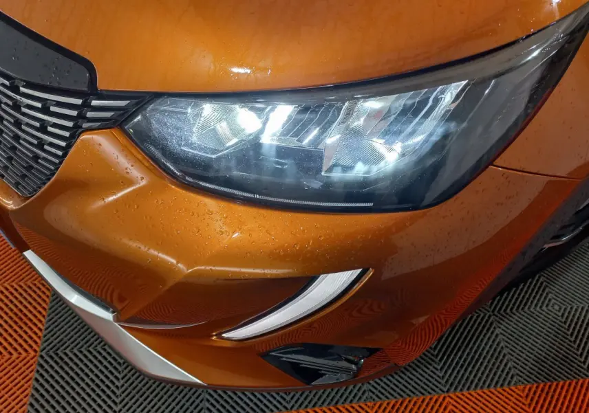Gros plan sur le phare avant droit et la calandre d’une Peugeot 2008 orange métallisé avec gouttes d’eau visibles.