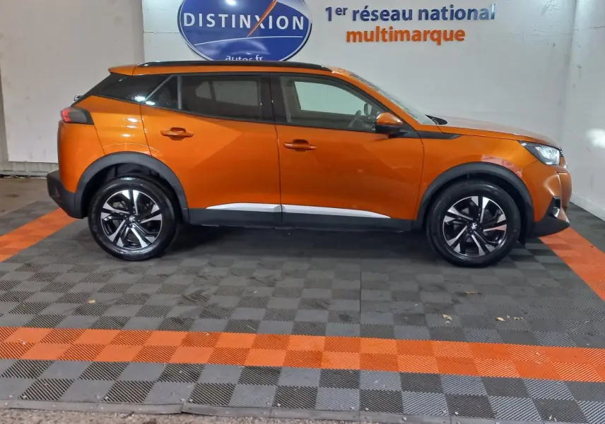 Profil droit d'un Peugeot 2008 orange 2021 avec jantes noires et toit noir dans un showroom multimarque.