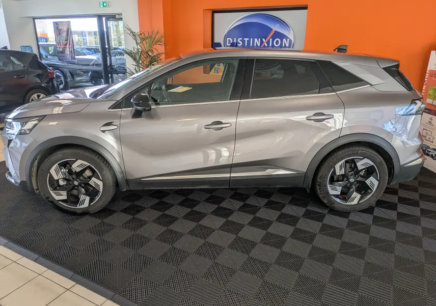 Profil droit du Renault Symbioz gris cassiopée métal 2025 en hybride essence, avec jantes noires et argentées en intérieur showroom.